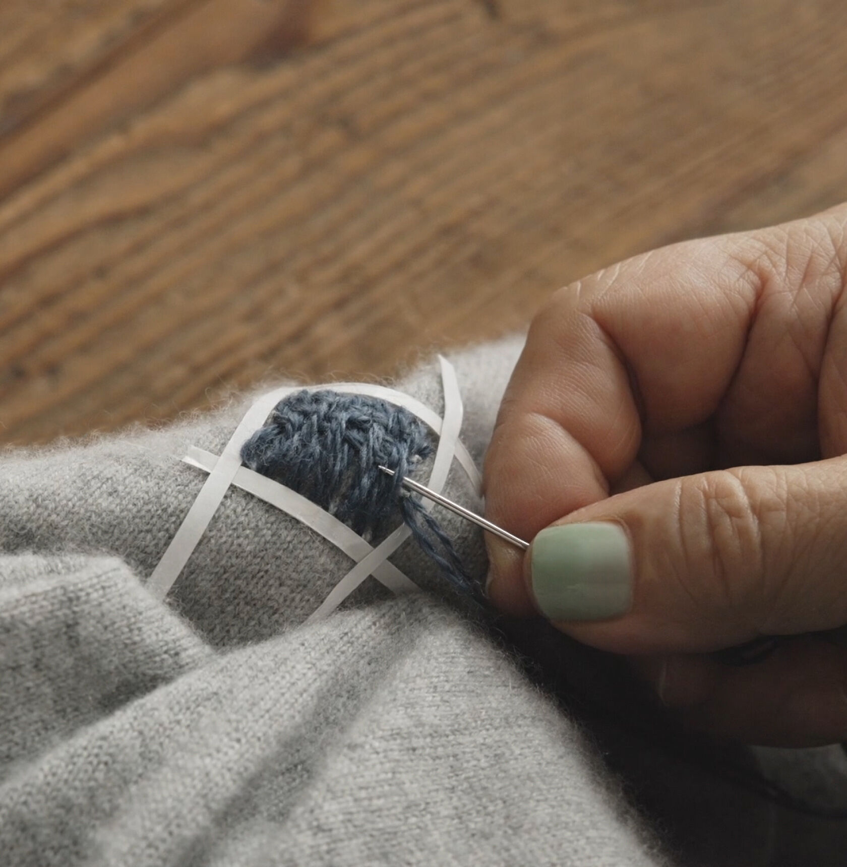 Close-up of hands weaving blue thread horizontally on a mended gray sweater.